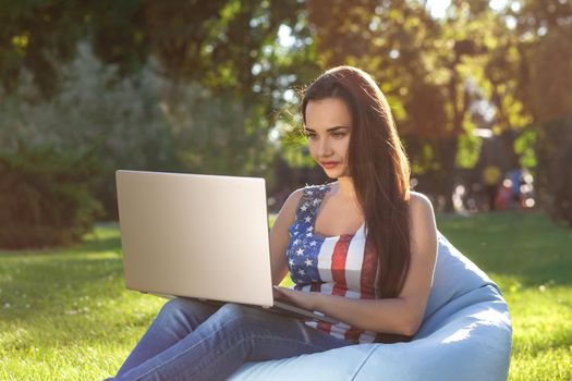 Young cute girl with laptop, sit on bean bag in garden or park, on green grass. Success small business, modern lifestyle, information technology, or online shopping concept