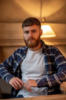 Portrait handsome bearded man wearing plaid shirt at modern cafe. Guy sitting in wooden chair and relaxing.