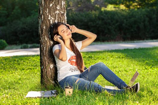 Woman relax with headphones listening to music sitting on grass in park. Young woman enjoys music and loneliness