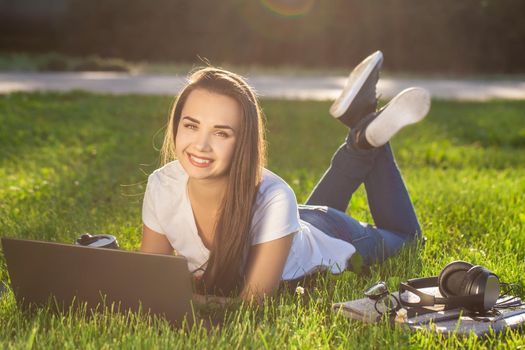 Young woman using laptop in the park lying on the green grass. Modern lifestyle, information technology or online shopping concept