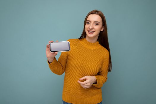 Photo shot of attractive positive young woman wearing orange sweater poising isolated on blue background with empty space holding in hand and showing mobile phone with empty space for mockup looking at camera.