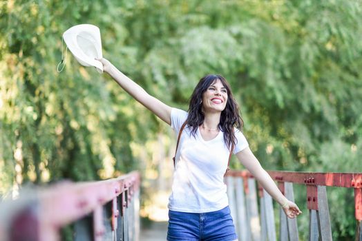 Beautiful hiker young woman with backpack, with blue eyes, wearing straw hat, hiking in the countryside and having fun on a rural bridge.