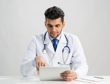 Young arab internist using tablet computer. Serious therapist in white medical gown with stethoscope sitting at desk. Modern digital technology in diagnosis and treatment. Medic data and healthcare