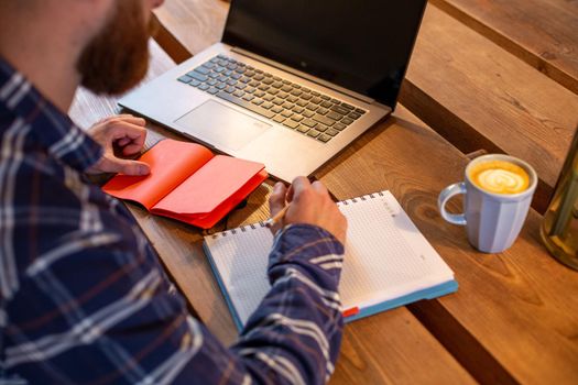 Cropped image of casual business man or freelancer planning his work on notebook, working on laptop computer with smart phone, cup of coffee on table at coffee shop or home office, working from cafe concept