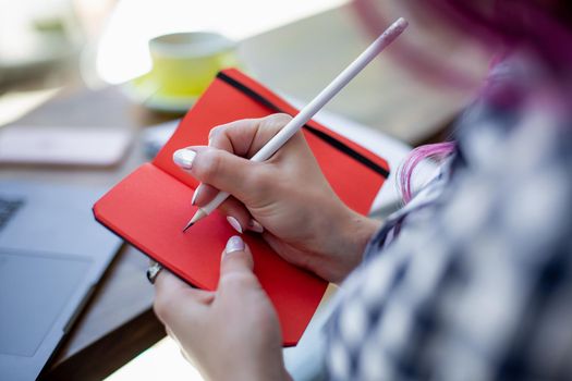 Positive female student with pink hair writing down funny story in notebook sitting in coffee shop with cup of cappuccino and laptop. Smiling female noting text in notepad
