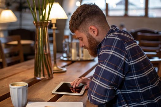Freelancer dressed in casual outfit focus on reading news and looking on digital tablet while sitting in cozy urban cafe. Young man using modern technology for search job vacancy in internet