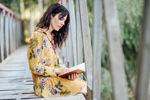 Young woman, wearing flowered dress, reading a book on a rural bridge.
