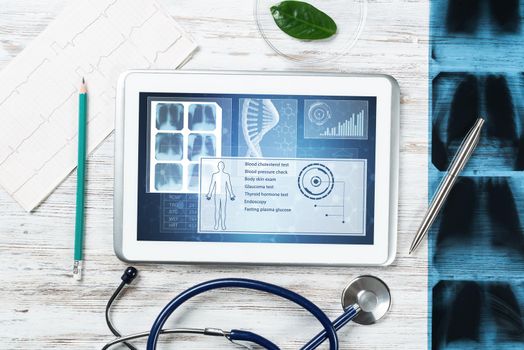 Modern social health insurance program. Tablet computer with healthcare application interface on screen. Stethoscope, x-ray image and cardiogram on wooden desk. Digital healthcare technologies.