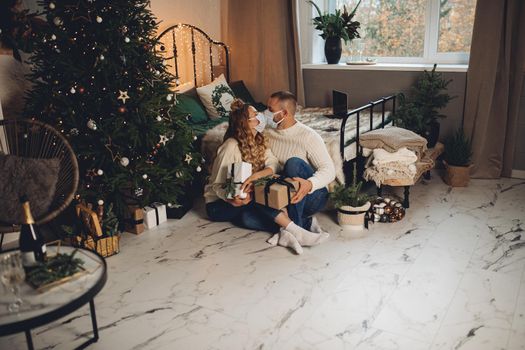 Kissing adult couple wearing face masks holding christmas presents sitting by bed and xmas tree.