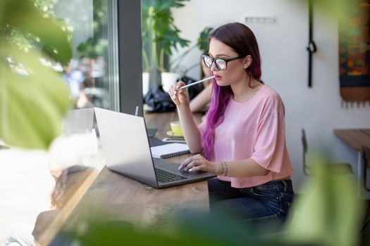 Side view of young businesswoman sitting at table in coffee shop. On table cup of coffee and laptop. In background white wall and window. Girl shopping online.