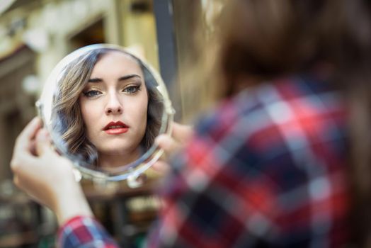 Portrait of young woman looking at herself in a little mirror in urban background.