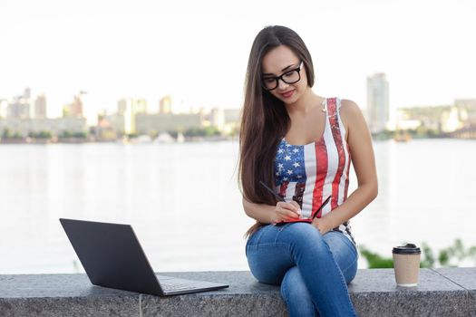 The beautiful young woman sits in the park near river with the laptop. Success small business, modern lifestyle, information technology, or online shopping concept