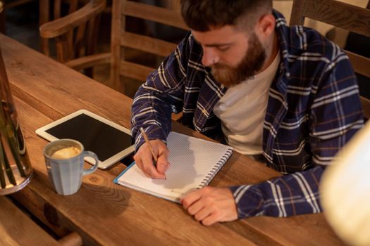 Young bearded businessman sits in cafe, home at table and writes in notebook, near lies tablet computer with black screen. Man is working, studying. Online education, marketing, training.