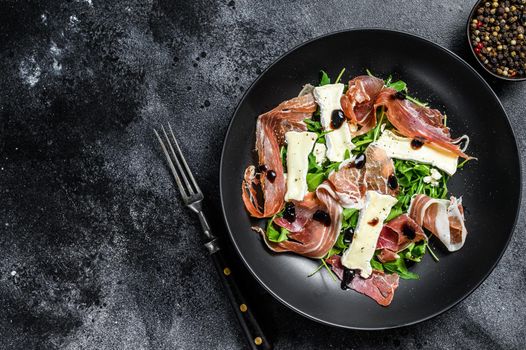 Prosciutto crudo ham salad with brie camembert cheese and arugula on a plate. Black background. Top view. Copy space.