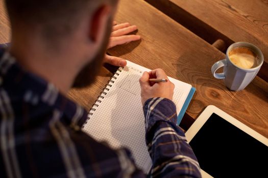 Young bearded businessman sits in cafe, home at table and writes in notebook, near lies tablet computer with black screen. Man is working, studying. Online education, marketing, training.
