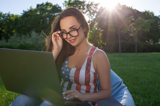 Pretty young woman sitting on bean bag use laptop while resting on grass in park on the sun. Success small business, modern lifestyle, information technology, or online shopping concept