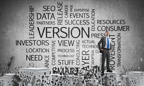 Confident businessman in suit standing on pile of documents among flying letters with business related terms on background. Mixed media.