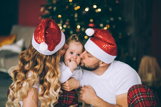 Loving mother and father in red Santa hats kissing sweet daughter. Christmas tree in background.