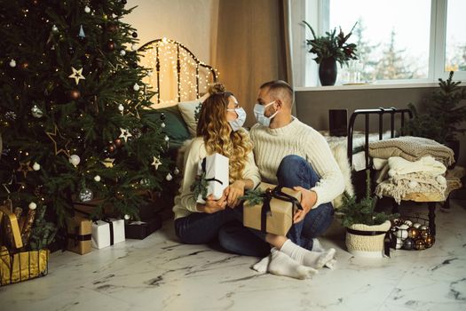 Young man and woman in masks looking each other while holding Christmas gifts in their bedroom