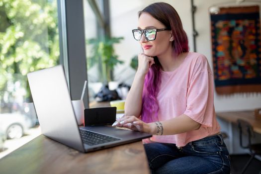 Side view of young businesswoman sitting at table in coffee shop. On table cup of coffee and laptop. In background white wall and window. Girl shopping online.