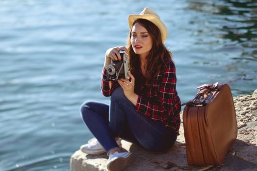 Young woman taking photographs with a vintage camera sitting in a harbour. Girl wearing plaid shirt, blue jeans and sun hat.