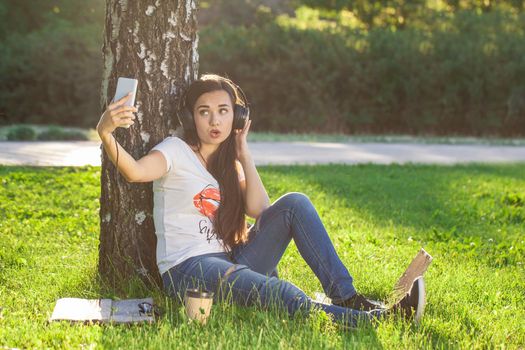 Woman relax with headphones listening to music sitting on grass in park. Young woman enjoys music and loneliness. Young woman with headphones doing selfie on her smartphone