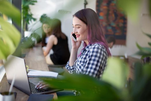 Side view of young businesswoman sitting at table in coffee shop. On table cup of coffee and laptop. In background white wall and window. Girl shopping online.