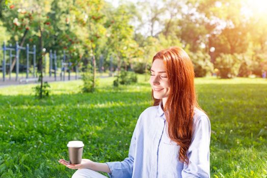 Young redhead woman meditating in lotus pose with cup of coffee on green grass. Beautiful calm girl in casual wear practicing of yoga with closed eyes in park. Mental relax and balance concept.