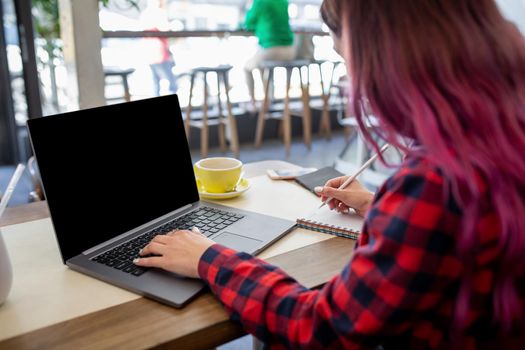 Rear shot of female student learning online via laptop before her lectures. Woman typing on notebook with blank screen with copy space for your text or advertising content, sitting at wooden table