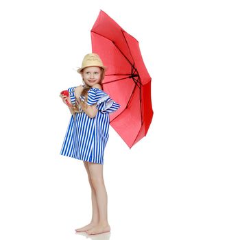 A stylish little girl with long blond hair in a very short, striped, summer blue dress and a straw hat.She is sheltered from the sun or rain under a red umbrella.