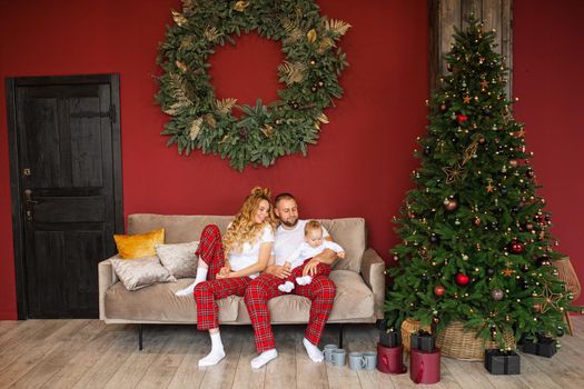 Cheerful Caucasian family in family look relaxing on cozy couch with baby in arms at Christmas.