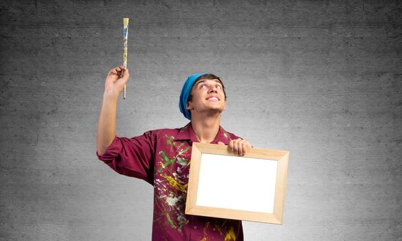 Young handsome painter artist holding paintbrush and blank canvas in frame. Portrait of happy painter on gray wall background. Creative hobby and artistic occupation. Art school advertising.