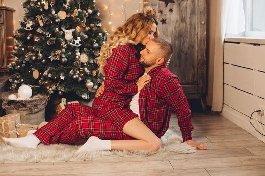 Pretty woman kissing her boyfriend while sitting on floor with Christmas tree on the background