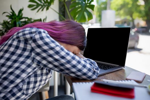 Young woman lying on her arms on the table in cafe in front of laptop with cup of coffee, sleepy. Rear shot of female student learning online via laptop before her lectures.