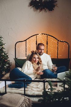 Couple in sweaters and jeans celebrating New Year with drinks and laptop on bed. Lockdown concept.