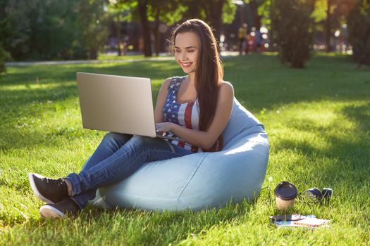 Young cute girl with laptop, sit on bean bag in garden or park, on green grass. Success small business, modern lifestyle, information technology, or online shopping concept
