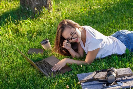 Young woman using laptop in the park lying on the green grass. Modern lifestyle, information technology or online shopping concept