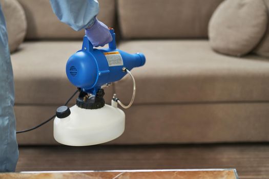 Close up of male hands in protective gloves using microparticle gun for virus prevention near sofa in hotel room. Coronavirus and quarantine concept