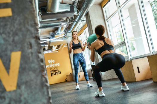 Rear view of a young athletic woman in sportswear squatting with weight ball at gym, working out with personal trainer. Sport and healthy lifestyle concept