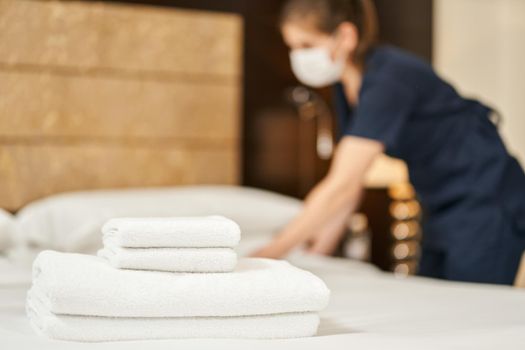 White fresh towels on the bed with a maid in a mask on the background. Hotel service concept