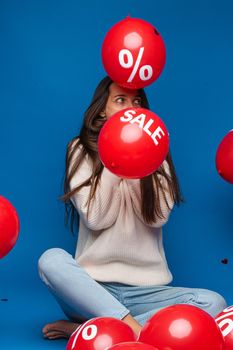 Portrait of shocked brunette woman in white sweater pulling her collar up to opened mouth and looking up. Red air balloons with sale and discount sign flying around.