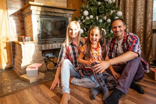 Theme Christmas and New Year family circle. Young Caucasian family sitting on wooden floor home in living room near fireplace Christmas tree.