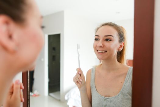 Young woman standing near mirror with toothbrush, wearing grey shirt. Concept of morning hygiene and healthy way of life.