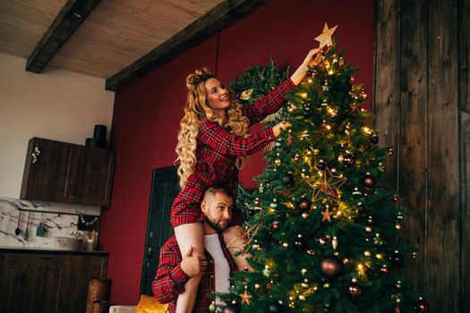 Smiling pretty lady decorating beautiful xmas tree with star while sitting on her husband shoulders