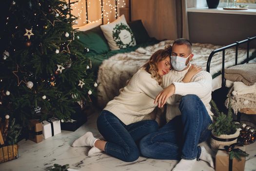 Portrait of loving wife hugging her husband wearing face mask. Sitting by bed and xmas tree.