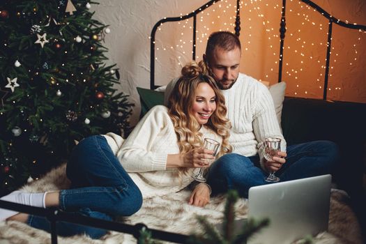 Happy young couple sitting on bed while holding glasses and making video call on laptop