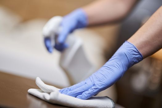 Cropped photo of maid cleaning and polishing wooden table with a spray detergent. Housekeeping and hygiene concept