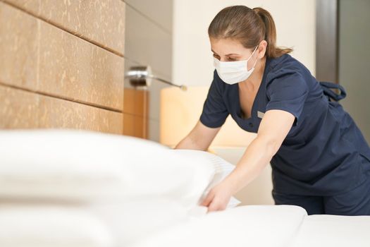 Maid wearing mask cleaning bedroom after guests in hotel room. Hotel service concept