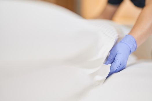 Close up of chambermaid in protective gloves fluffing white pillow on bed while cleaning in bedroom. Hotel service concept