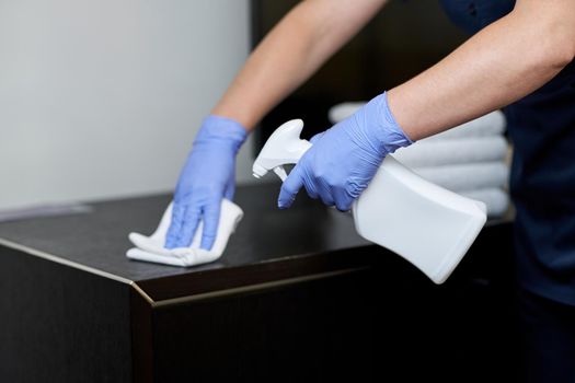 Female hands in protective gloves spraying disinfectant on furniture in hotel room. Hotel service concept